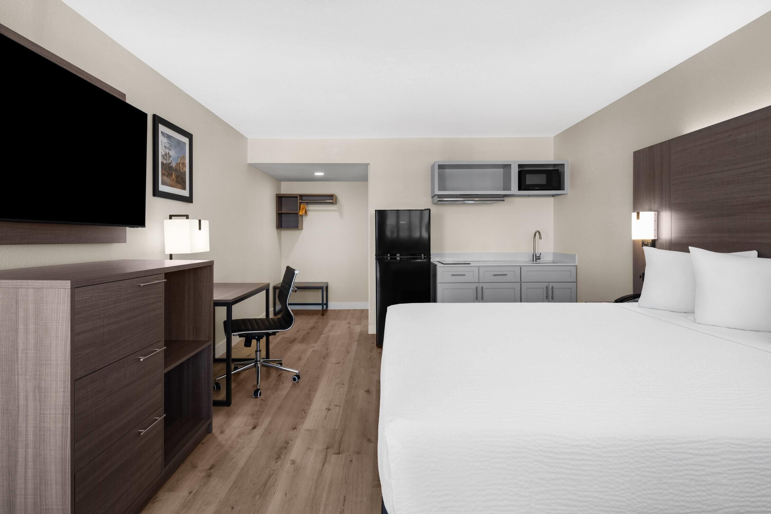 A hotel room with a bed, a desk, a chair, a TV, a microwave, and a kitchenette.