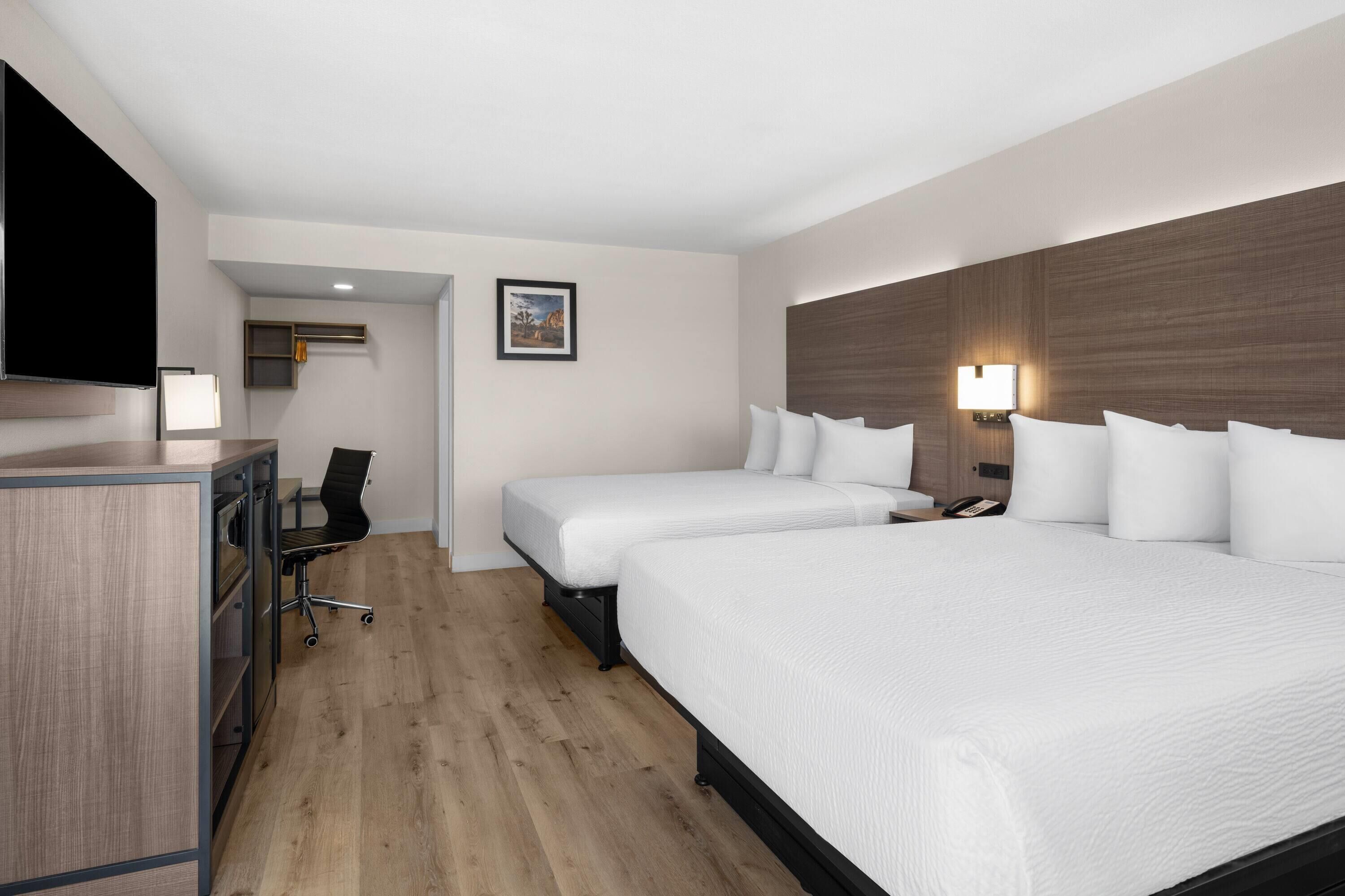A hotel room with a desk, two beds, a chair, a TV, and a wall-mounted lamp.