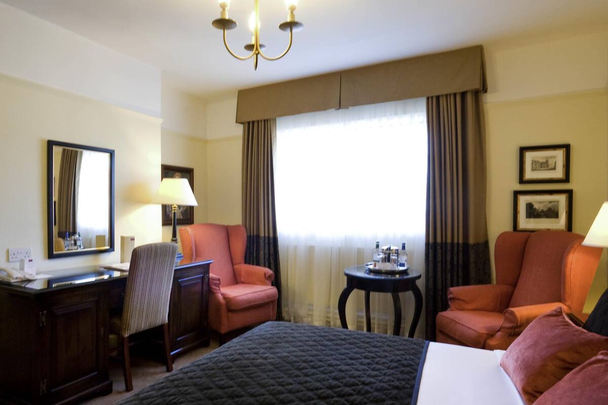 A hotel room with a bed, two armchairs, a desk, a chair, a small table with bottles, a lamp, a mirror, and curtains.