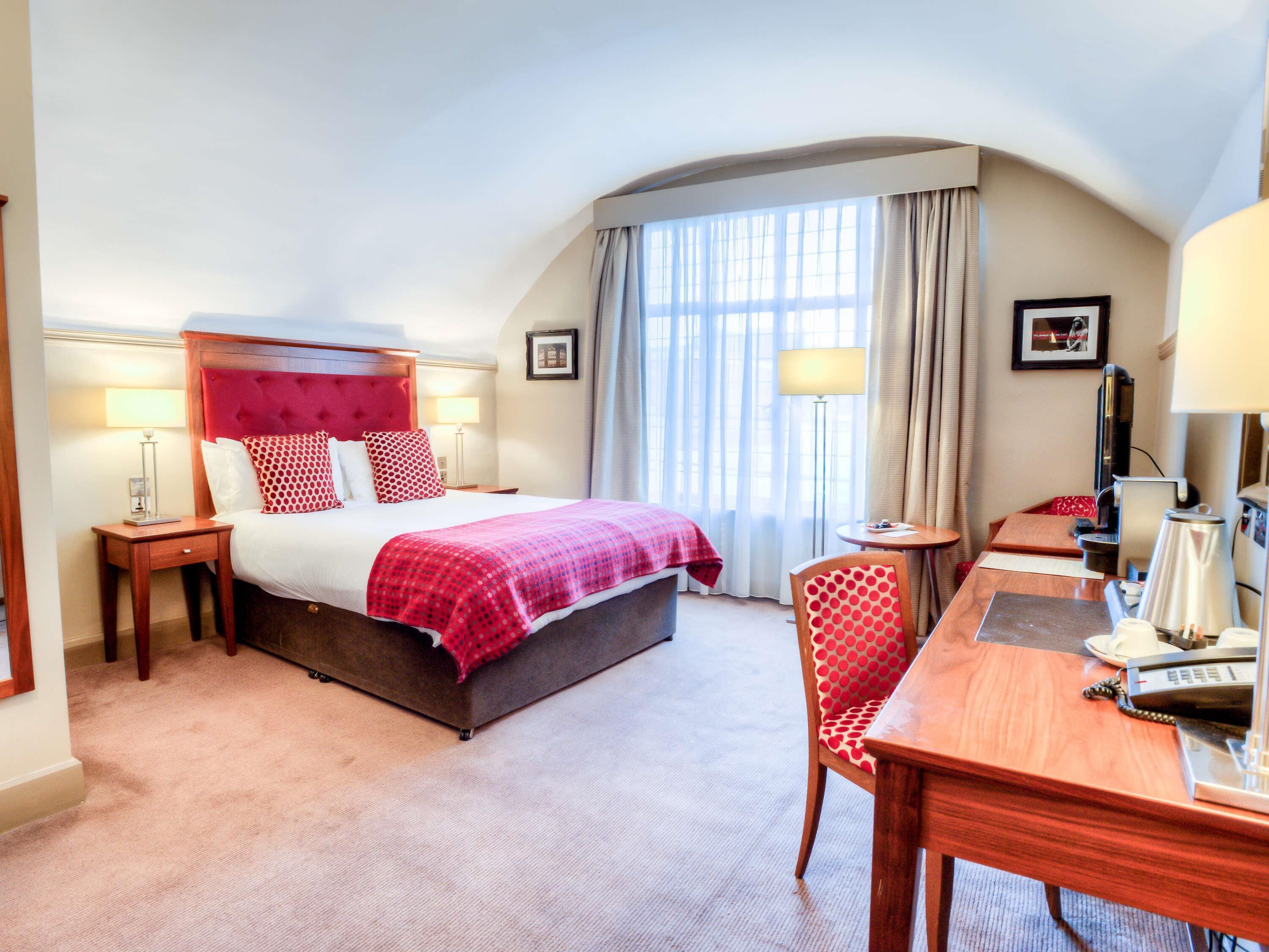 Une chambre d’hôtel avec un grand lit, un bureau, une chaise, une télévision et une fenêtre avec des rideaux.