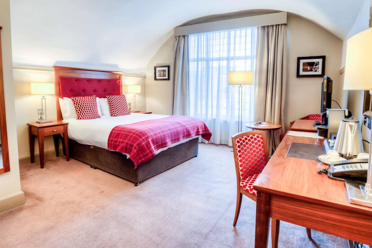 A hotel room with a large bed, a desk, a chair, a TV, and a window with curtains.