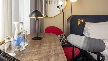 A hotel room with a wooden table, a desk with a computer, a bed with a headboard, and a mirror on the wall.