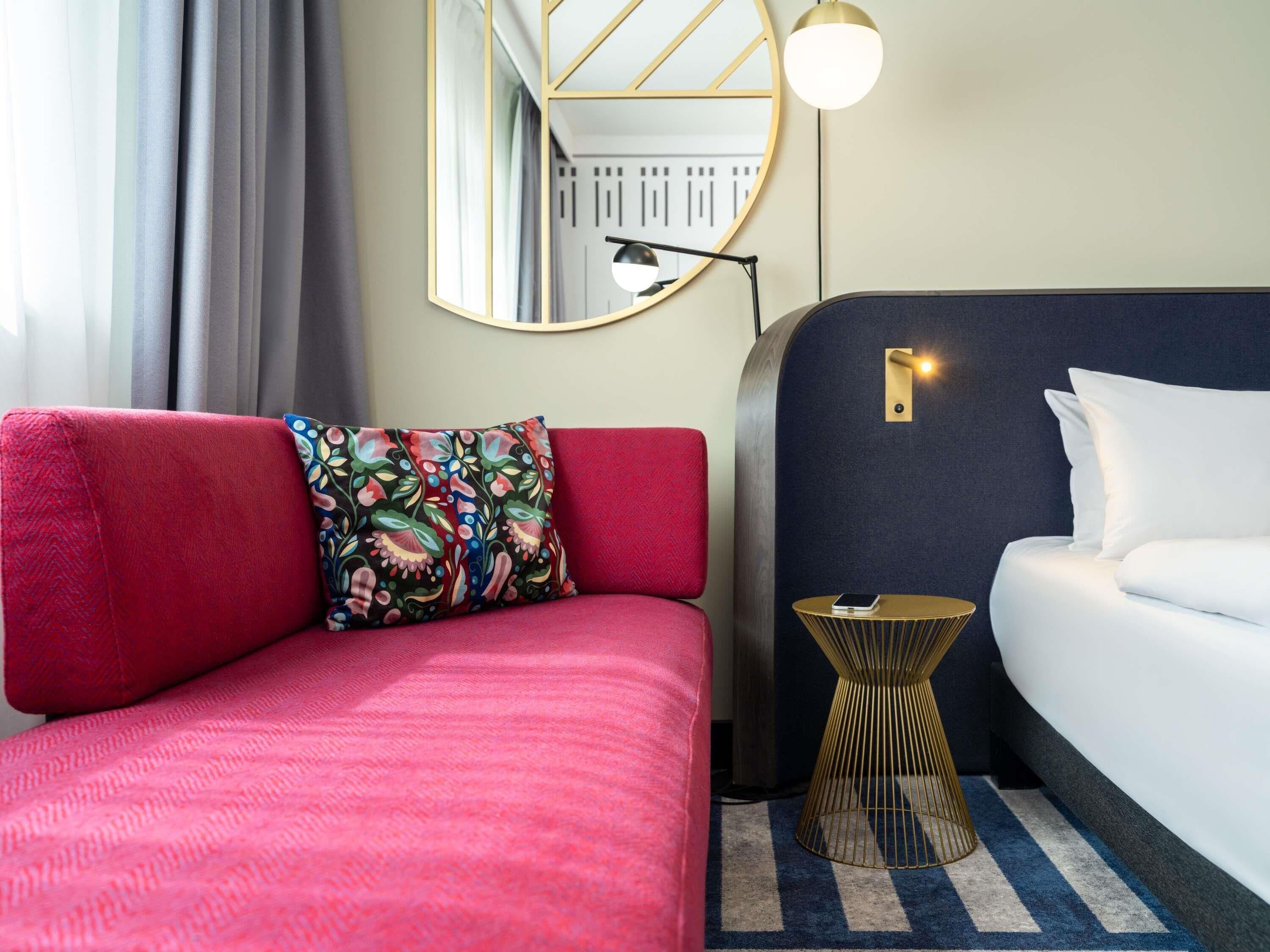 Ein Hotelzimmer mit rotem Sofa, einem gemusterten Kissen, einem runden Spiegel, einem Nachttisch und einem Bett mit weißer Bettwäsche.