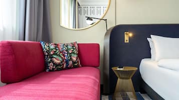 Ein Hotelzimmer mit rotem Sofa, einem gemusterten Kissen, einem runden Spiegel, einem Nachttisch und einem Bett mit weißer Bettwäsche.