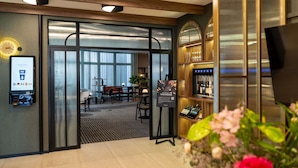 Un hall d’hôtel comprenant un comptoir d’accueil, un espace salon avec des tables et des chaises, un bar avec des bouteilles exposées, et une grande fenêtre avec des rideaux.
