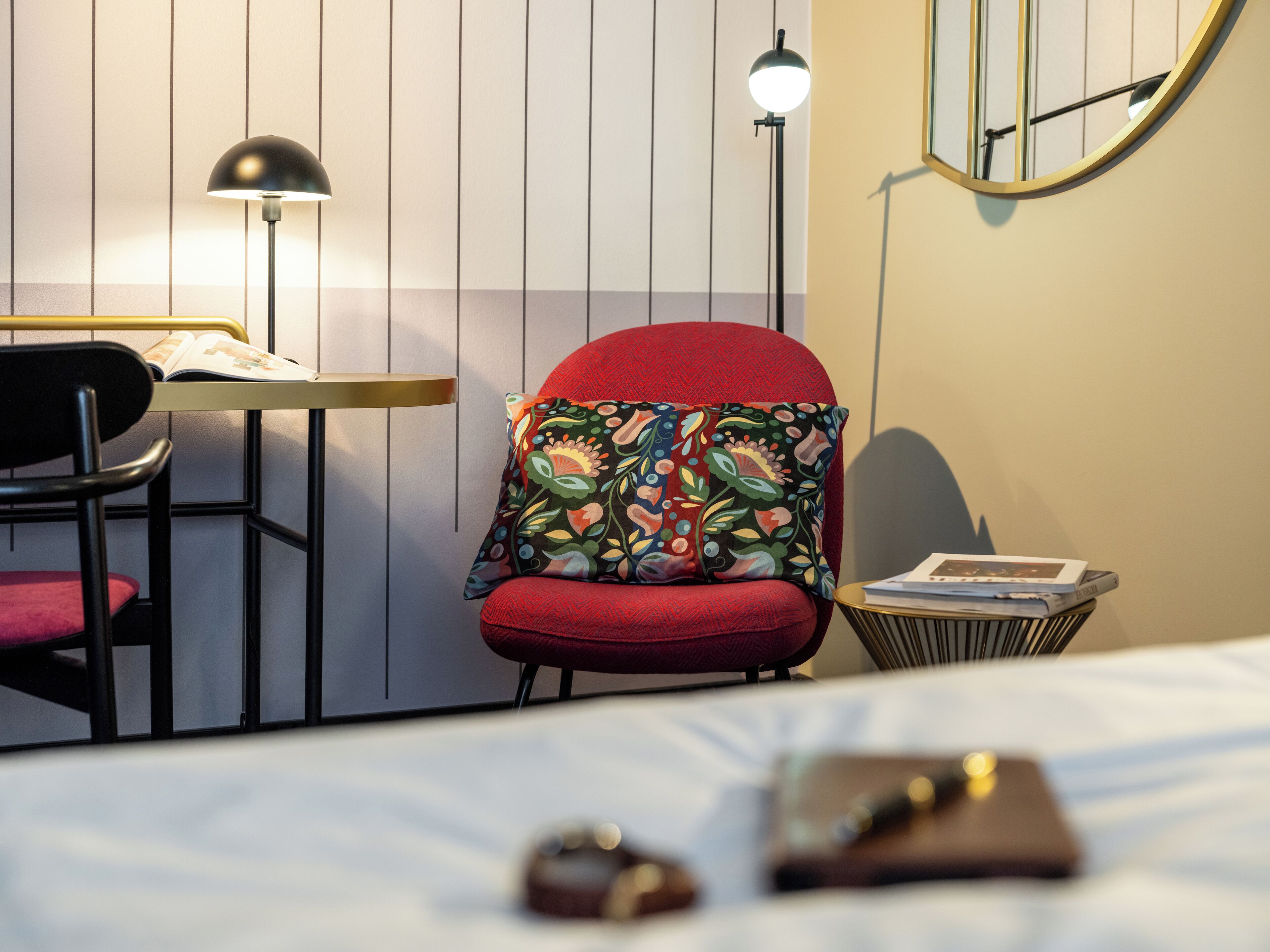 A hotel room with a bed, a red armchair with a colorful cushion, a desk with a lamp, and a small side table with books.