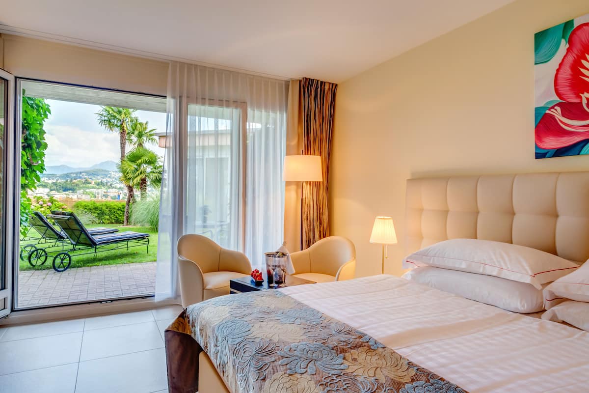 Habitación de hotel con una cama grande, dos sillones, una mesita, una lámpara y vistas a un jardín y montañas desde la ventana.