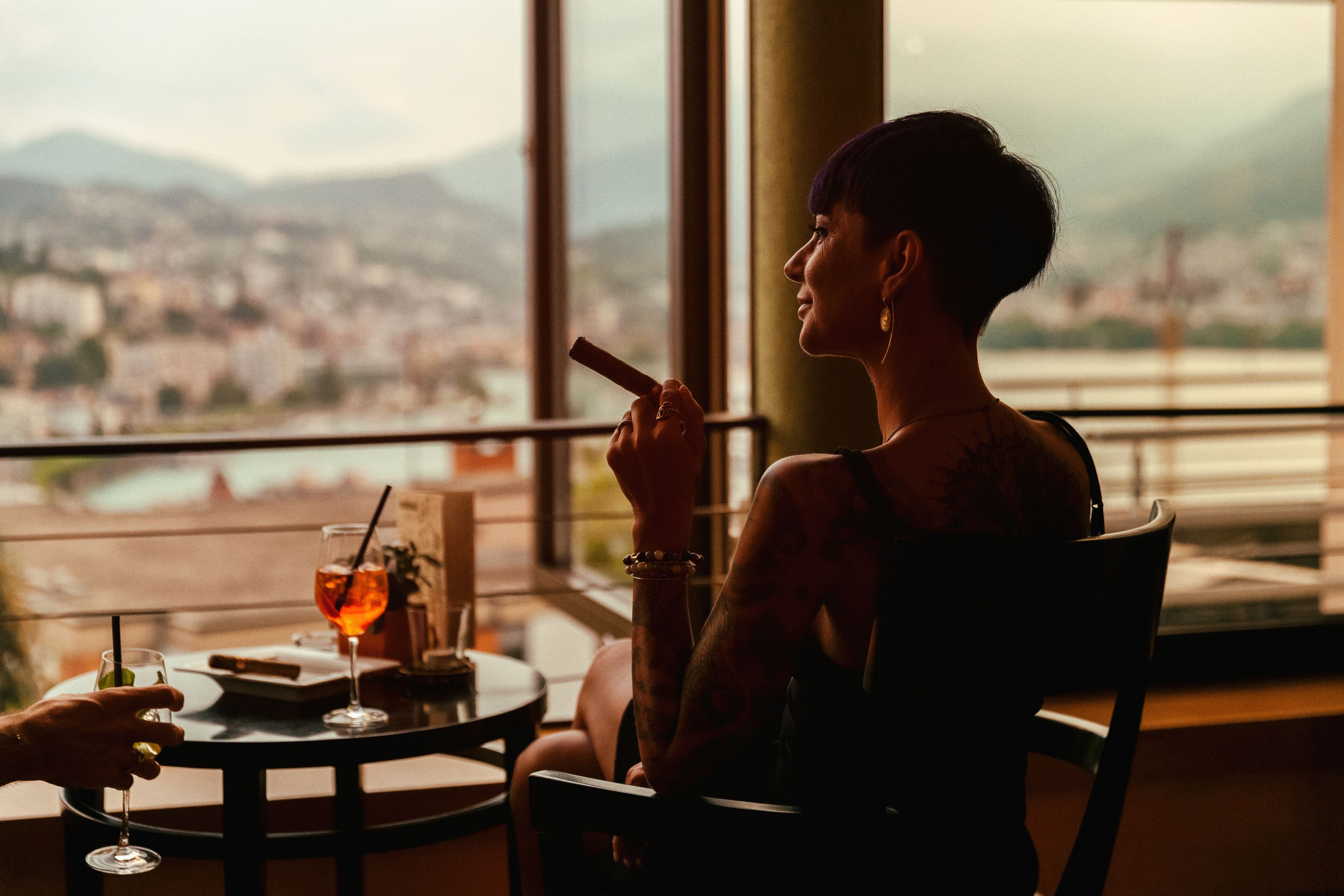 A person seated at a table with a drink, overlooking a cityscape through large windows.