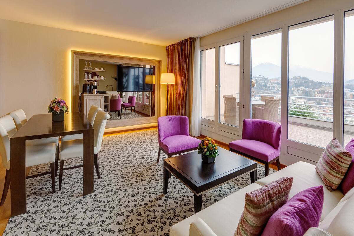 Habitación de hotel moderna con comedor, dos sillones morados, un sofá con cojines estampados y un ventanal con vistas a la ciudad.
