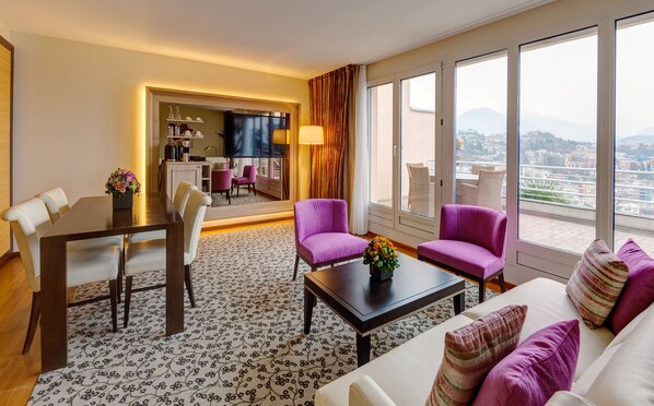 Habitación de hotel moderna con comedor, dos sillones morados, un sofá con cojines estampados y un ventanal con vistas a la ciudad.