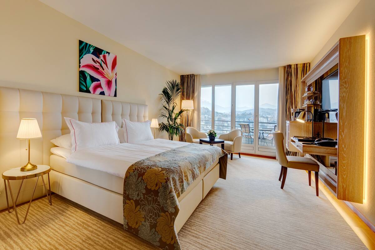Habitación de hotel con una cama grande, un escritorio, una silla y vistas a la ciudad a través de amplios ventanales.