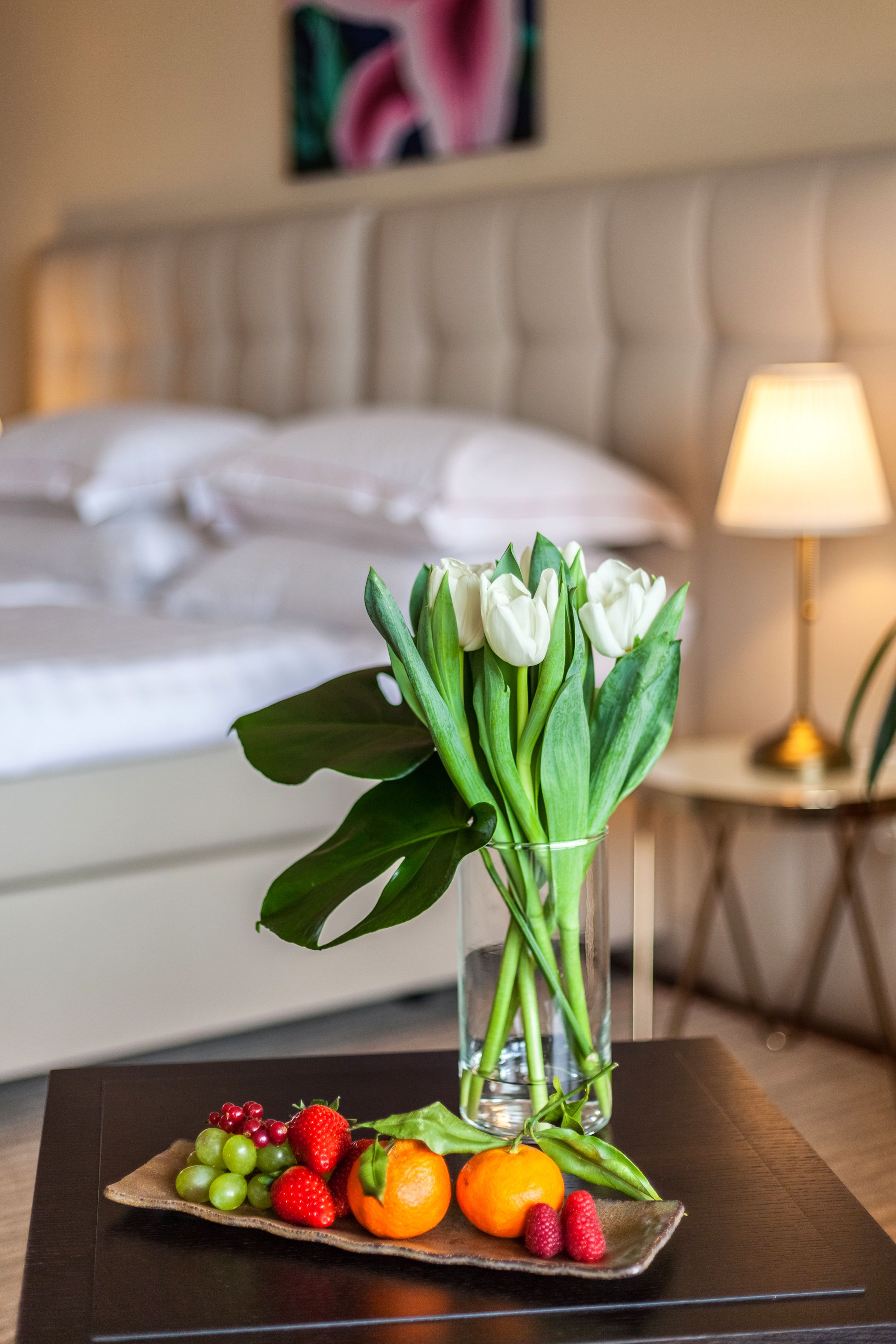 Una presentación ordenada de frutas y flores sobre una mesa en una habitación de hotel, con una cama y una lámpara al fondo.