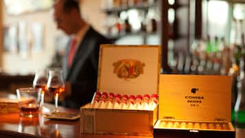 Un bar con una caja de cigarros Cohiba BeHike, vasos de whisky y un hombre al fondo.