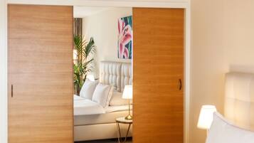 A hotel room with a bed, a bedside table with a lamp, a white sofa, a potted plant, and a painting on the wall.