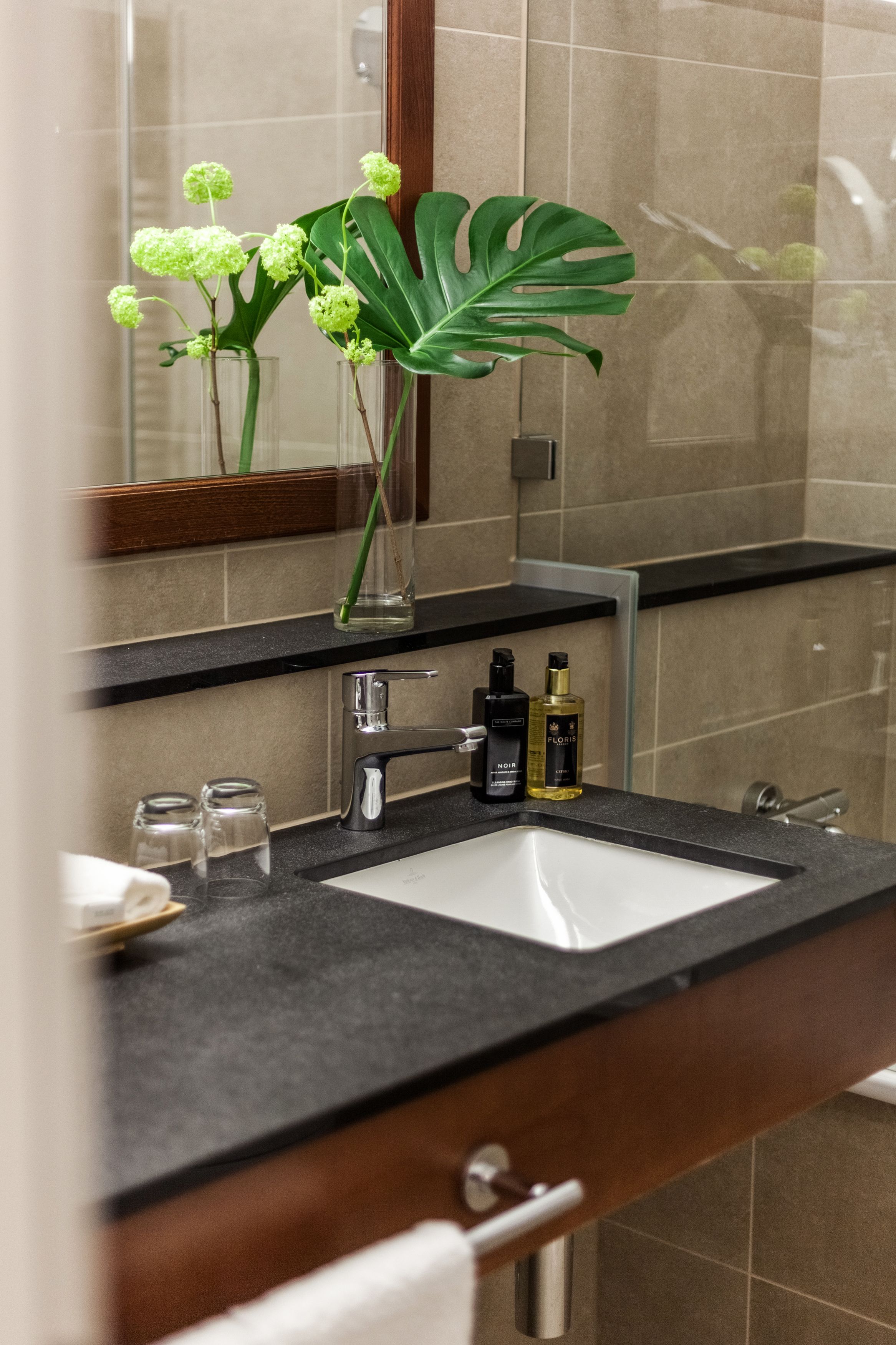 A bathroom with a black countertop, a white sink, and a mirror. There are two bottles on the counter, a glass, and a plant with green leaves.