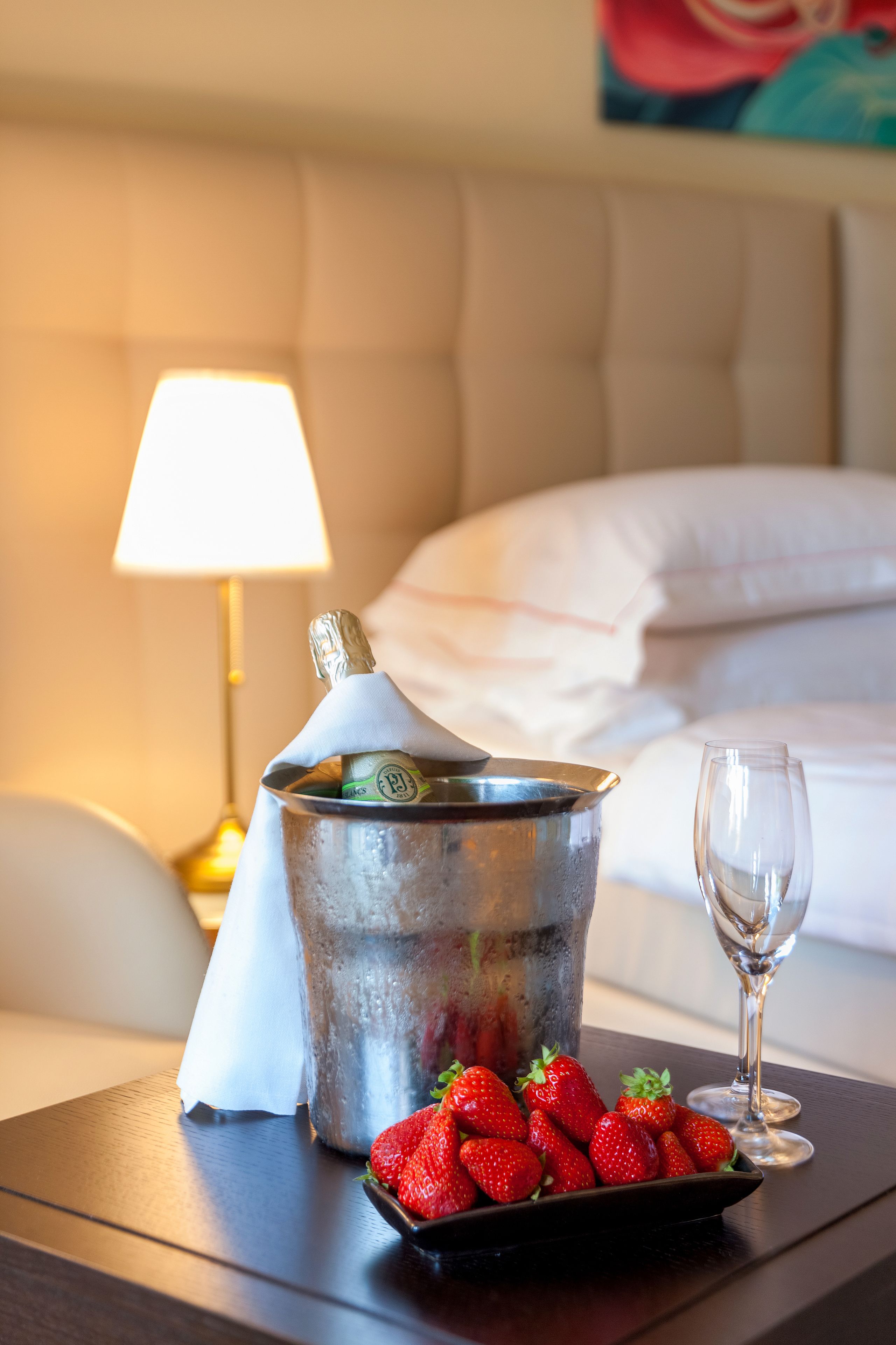 Un lit recouvert de draps blancs, une lampe de chevet, un seau contenant une bouteille de champagne, un verre et une assiette de fraises sur une table de chevet en bois.