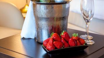 Un lit recouvert de draps blancs, une lampe de chevet, un seau contenant une bouteille de champagne, un verre et une assiette de fraises sur une table de chevet en bois.