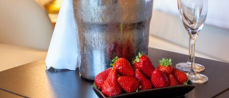 A bed with white linens, a bedside lamp, a bucket with a bottle of champagne, a glass, and a plate of strawberries on a wooden nightstand.