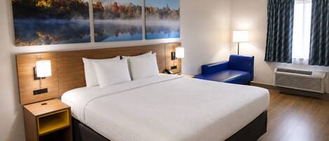 A hotel room with a large bed, a nightstand, a blue chair, a wall-mounted lamp, and a large window with curtains.