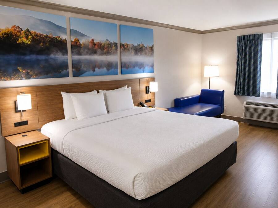 Habitación de hotel con una cama grande, una mesita de noche, una silla azul, una lámpara de pared y un ventanal con cortinas.