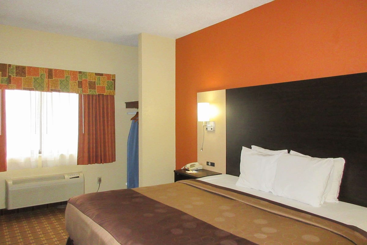 Quarto de hotel com cama, mesa de cabeceira, luminária e janela com cortinas.