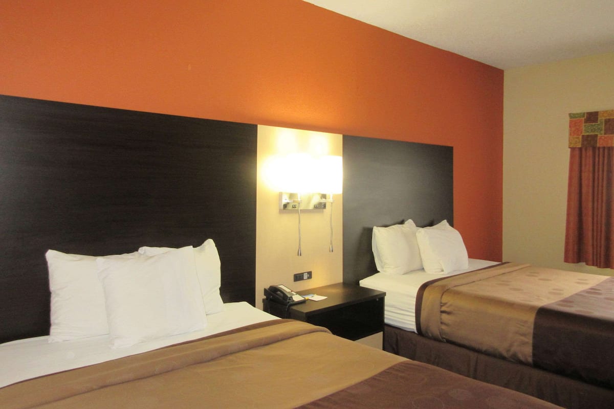 Quarto de hotel com duas camas, uma mesa de cabeceira com abajur e uma luminária na parede.