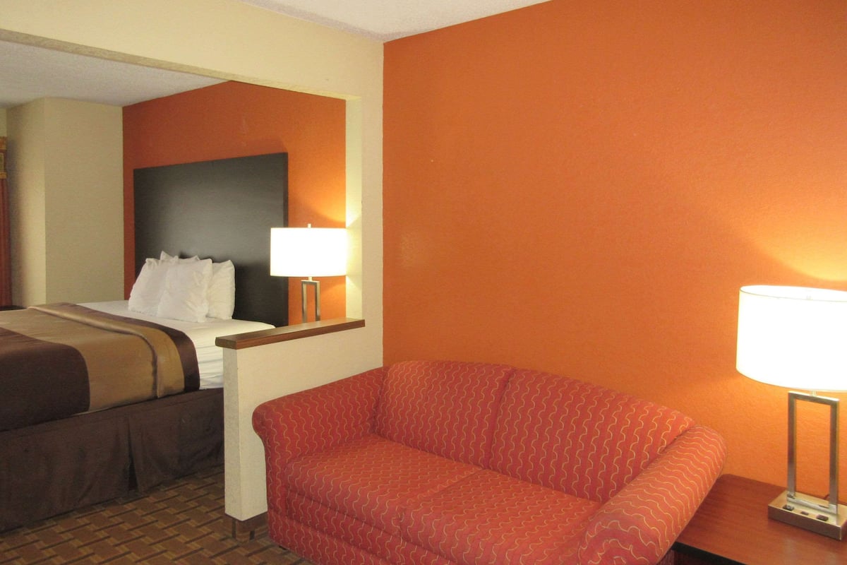 Quarto de hotel com cama, sofá vermelho, mesa de cabeceira com luminária e espelho fixado na parede.