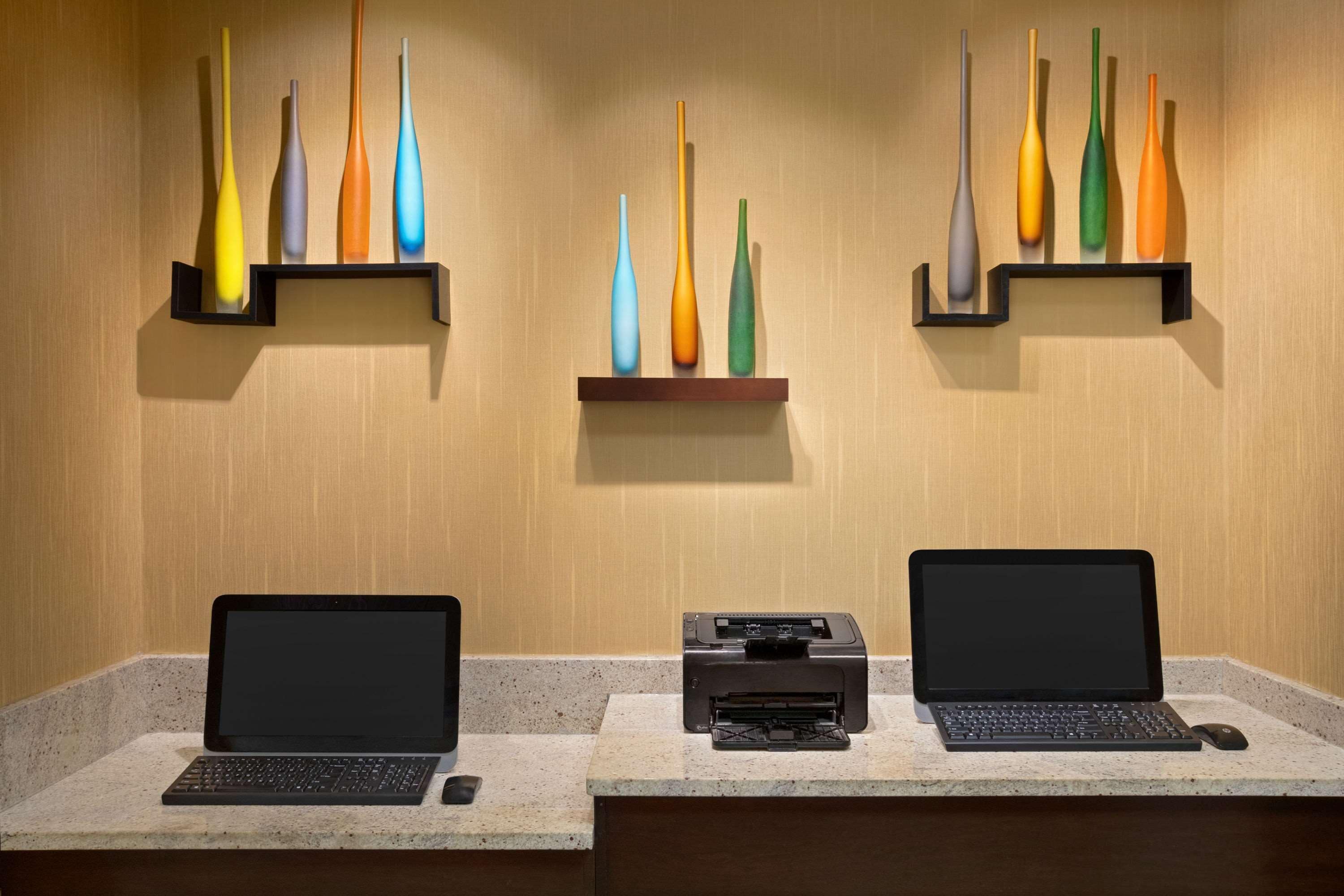 A desk with two laptops and a printer, and three shelves with colorful vases against a wooden wall.