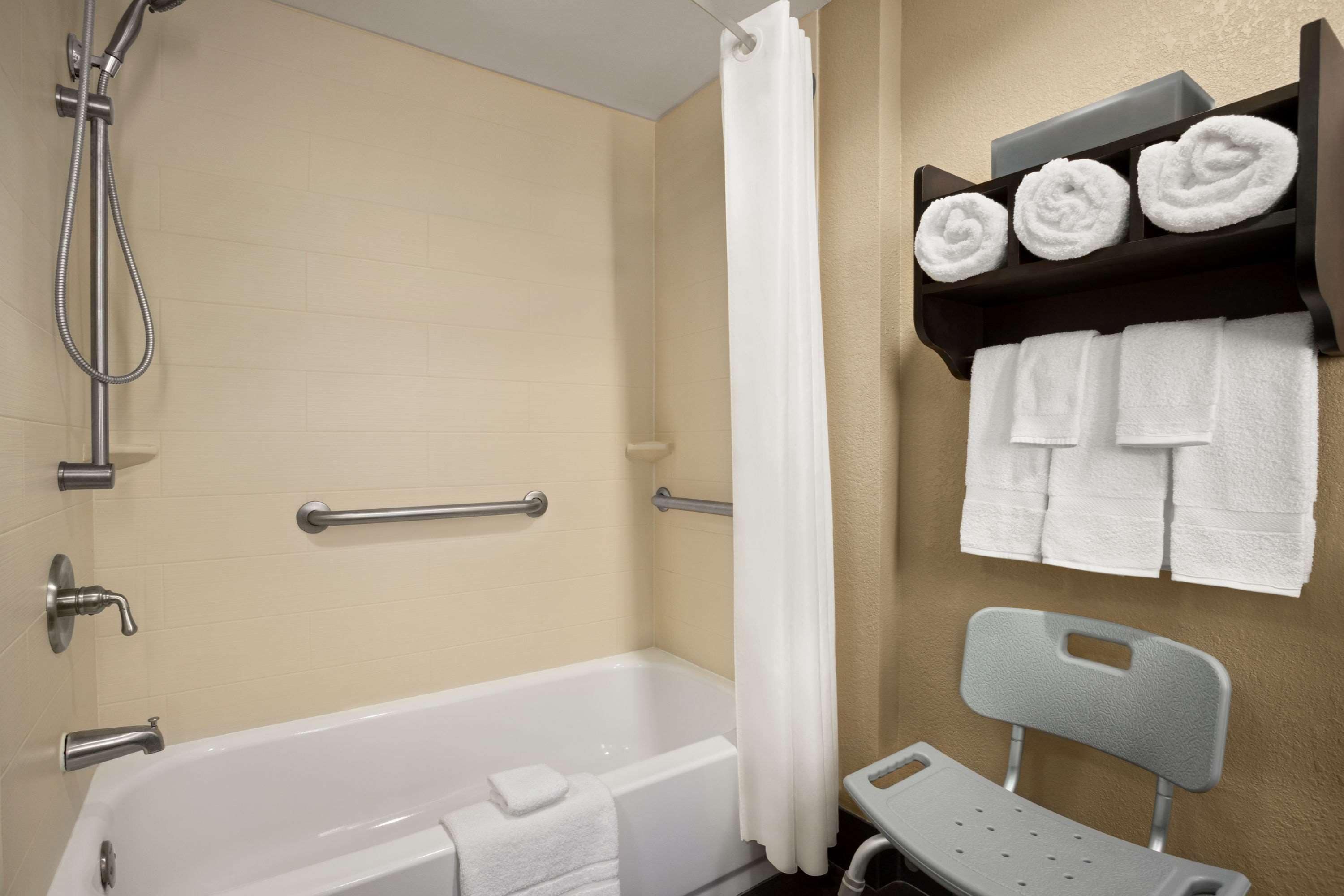 A bathroom with a bathtub, shower, grab bars, a towel rack with towels, and a chair.
