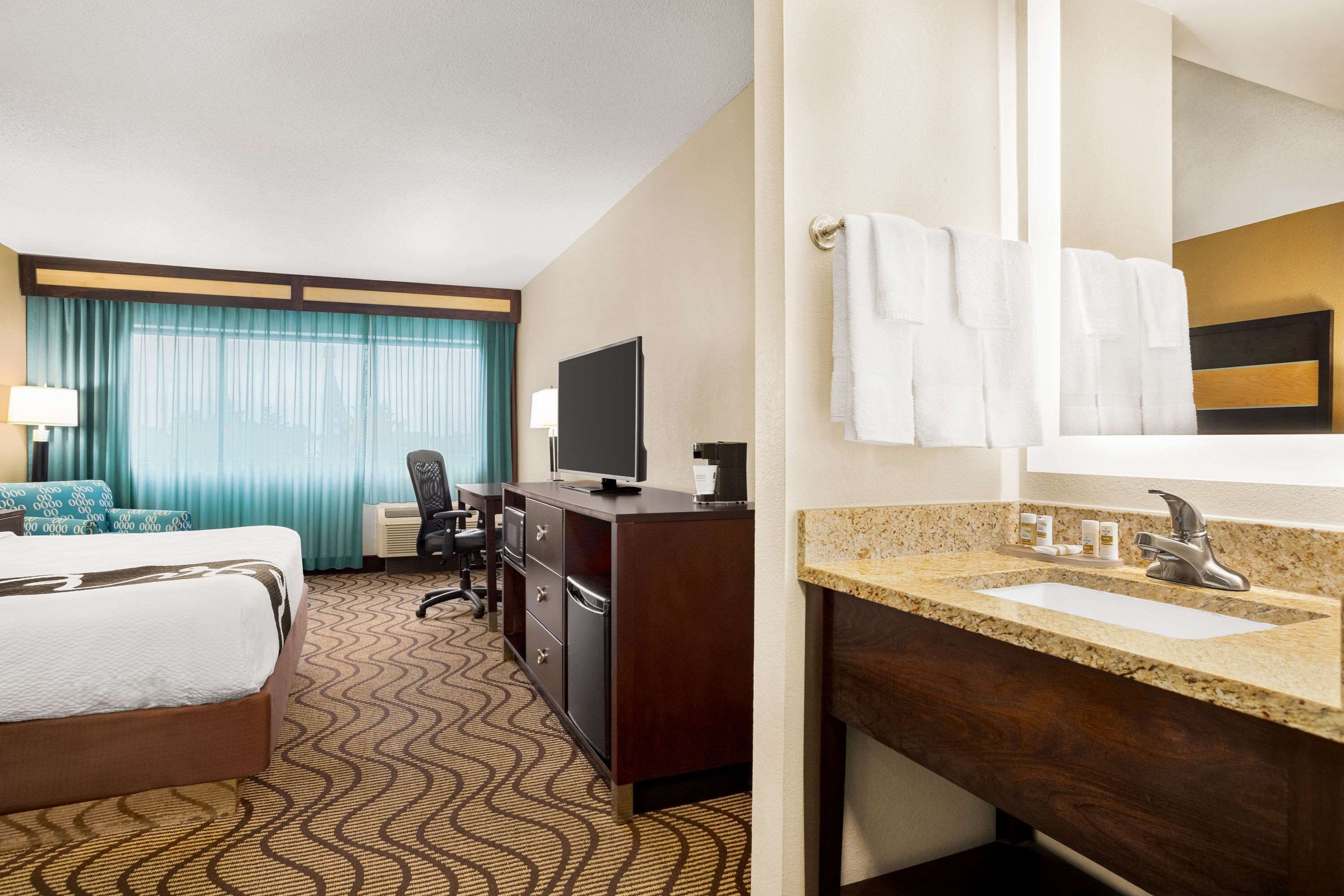 A hotel room with a bed, a desk with a computer, a TV, a bathroom with a sink and mirror, and a window with curtains.