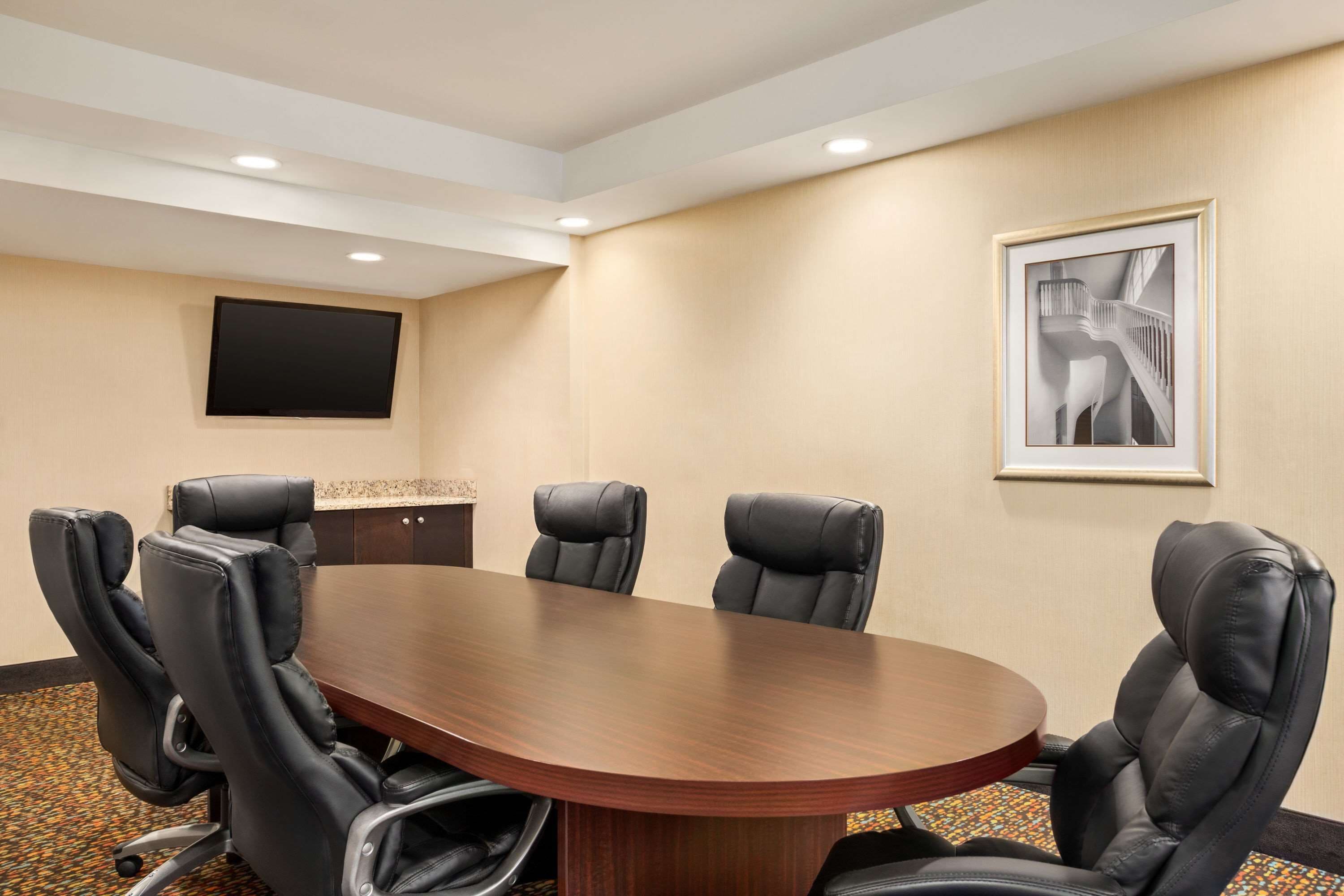 Uma sala de conferências com uma grande mesa oval, seis cadeiras de couro preto, uma TV de tela plana na parede e um quadro decorativo.