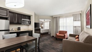 A hotel room with a kitchenette, a dining area, a living area with a sofa and armchair, and a bed with a window view.