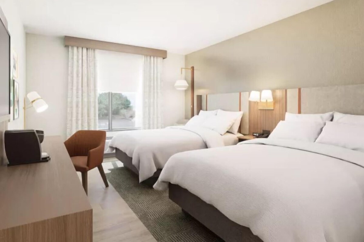 A hotel room with two beds, a desk, a chair, and a large window.