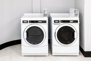 Twee witte wasmachines met voorlaadopening en digitale displays, naast elkaar geplaatst in een ruimte met betegelde vloer en een witte muur.