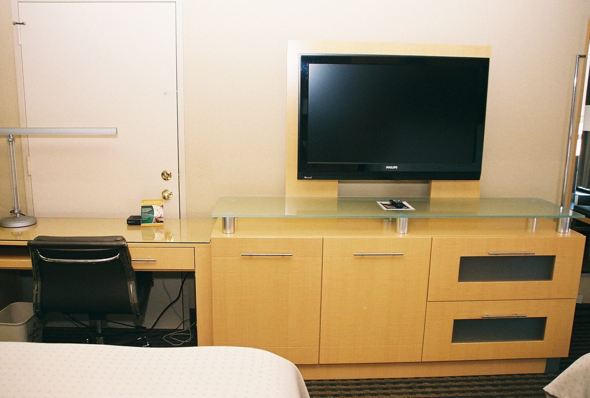 A hotel room with a bed, a desk, a television on a glass shelf, a chair, and a lamp.