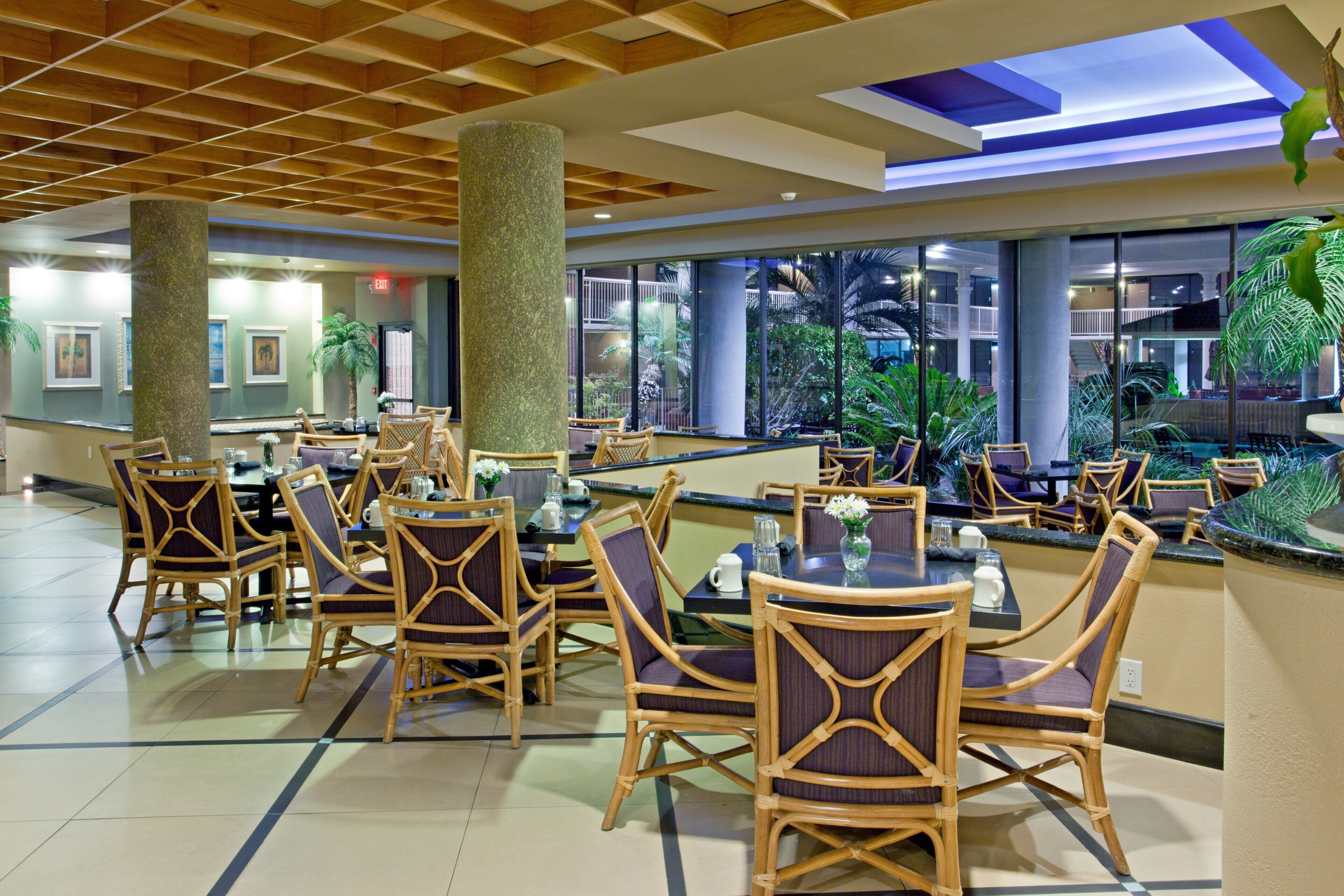 A hotel lobby with a seating area featuring wicker chairs and tables, a central column, and a view of a landscaped outdoor area.