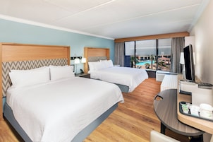 Quarto de hotel com duas camas, uma escrivaninha com cadeira, televisão e vista para a piscina.