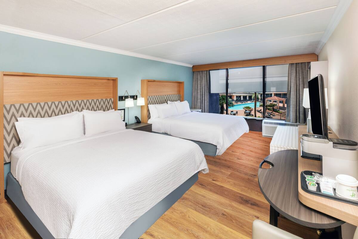 A hotel room with two beds, a desk with a coffee maker, a TV, and a view of a pool and outdoor area.