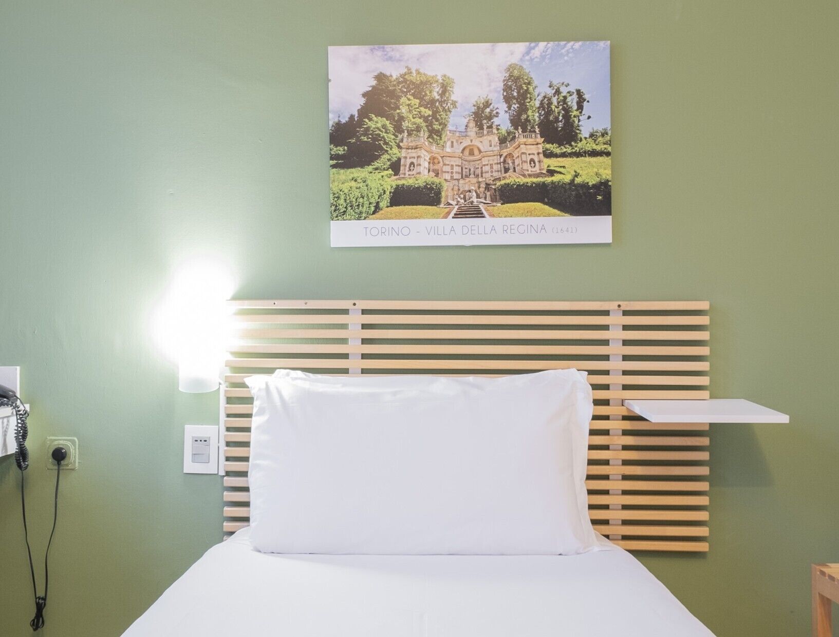 Una camera da letto con una testiera in legno, un cuscino bianco, un quadro con una villa e un comodino.
