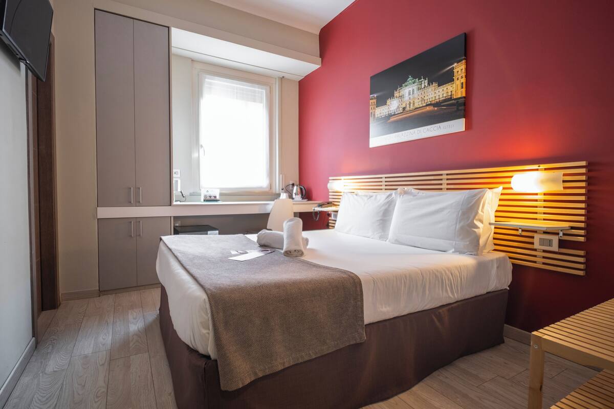 Une chambre d’hôtel avec un lit, un bureau, une chaise, une fenêtre et un tableau au mur.