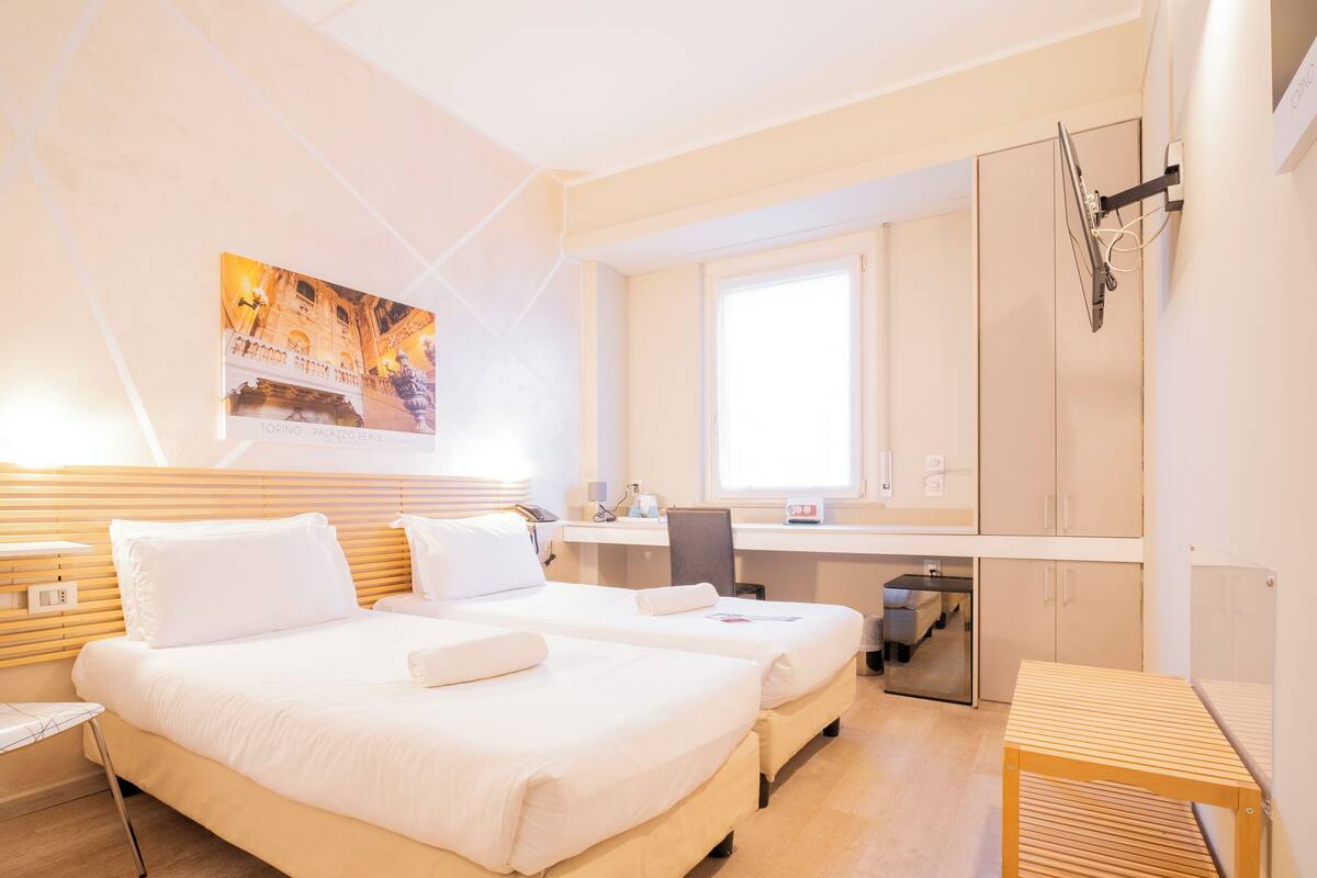 Une chambre d’hôtel avec deux lits, un bureau, une chaise, une petite table, une fenêtre et un tableau au mur.