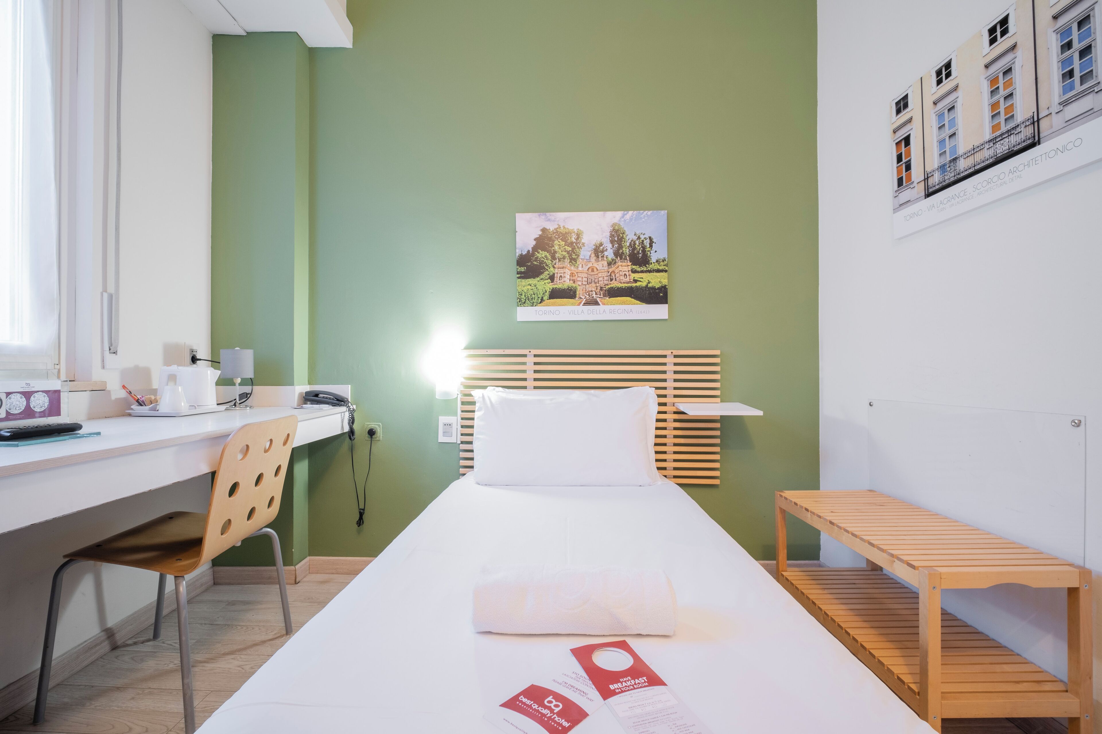 Ein Hotelzimmer mit einem Bett, einem Schreibtisch, einem Stuhl, einer Holzbank und einem an der Wand hängenden Bild.