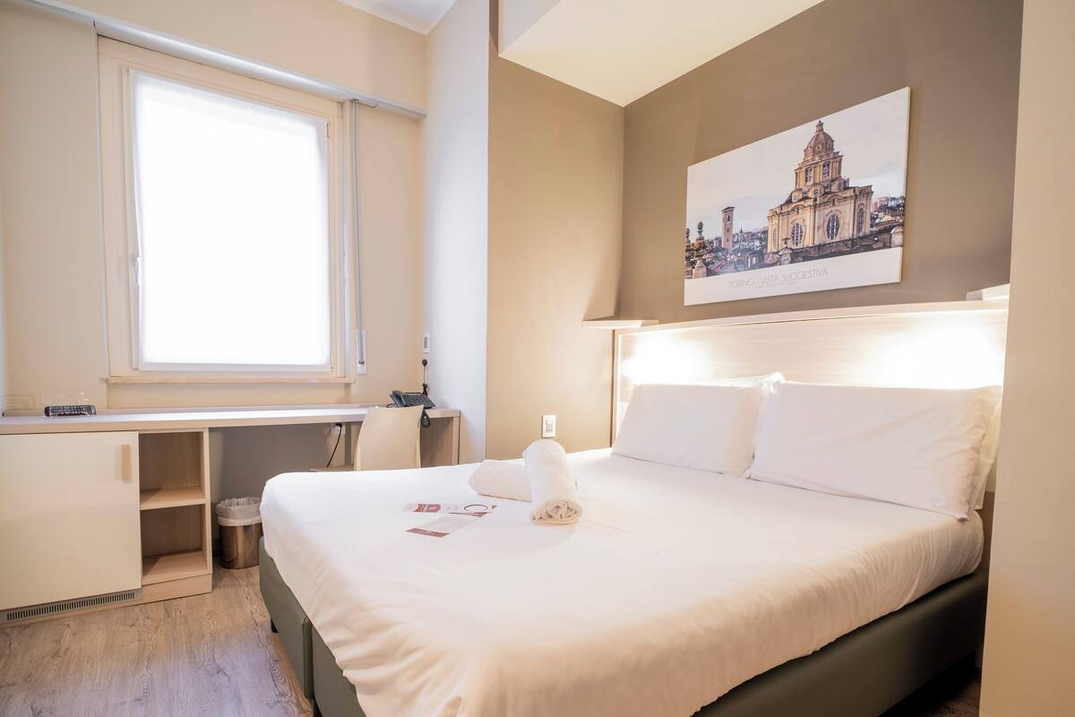 Une chambre d’hôtel avec un lit, un bureau, une chaise, une fenêtre et un tableau au mur.