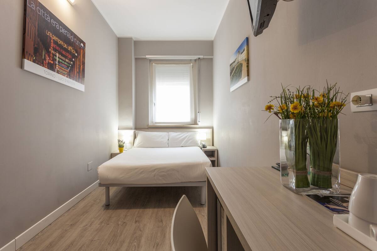 Une chambre d’hôtel comprenant un lit, une table de chevet, une commode, une fenêtre avec stores et un vase de fleurs.