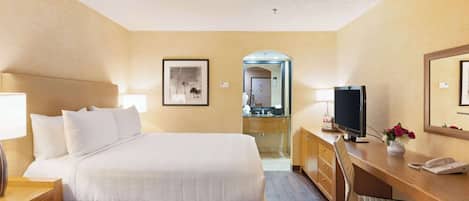 Habitación de hotel con cama, escritorio, silla, televisión y un baño visible a través de un arco.