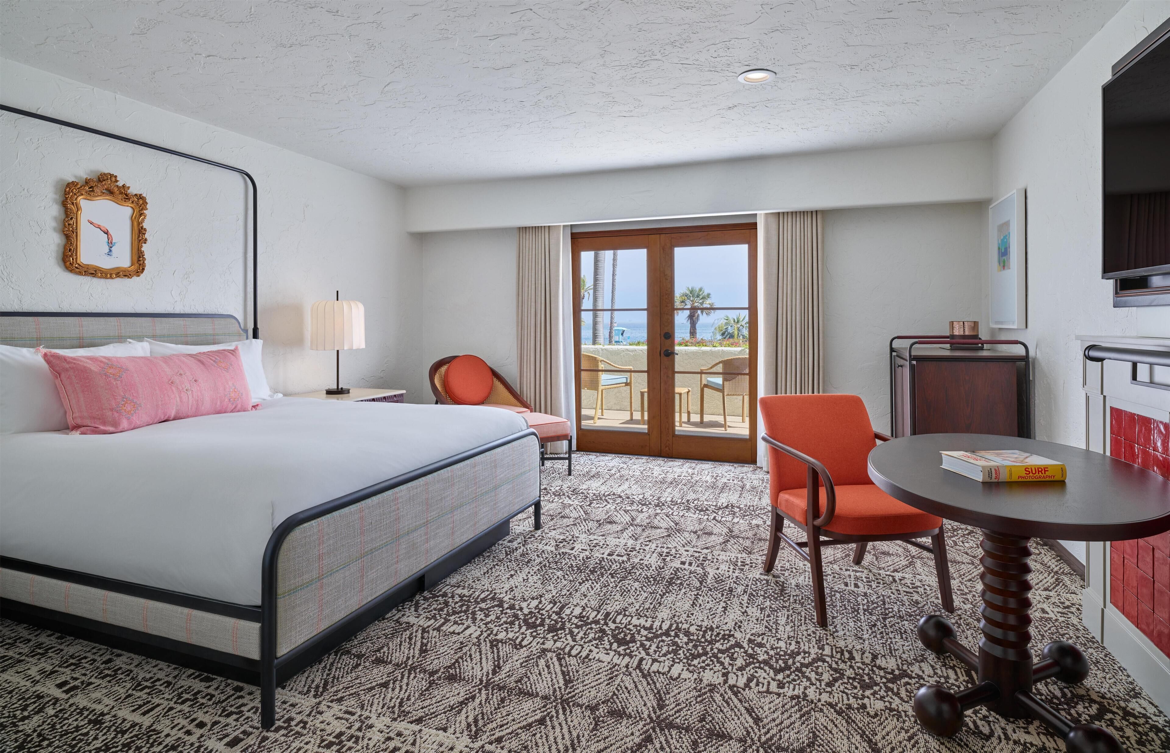 Habitación de hotel con cama, una silla, una mesa y vista a la playa por la puerta.