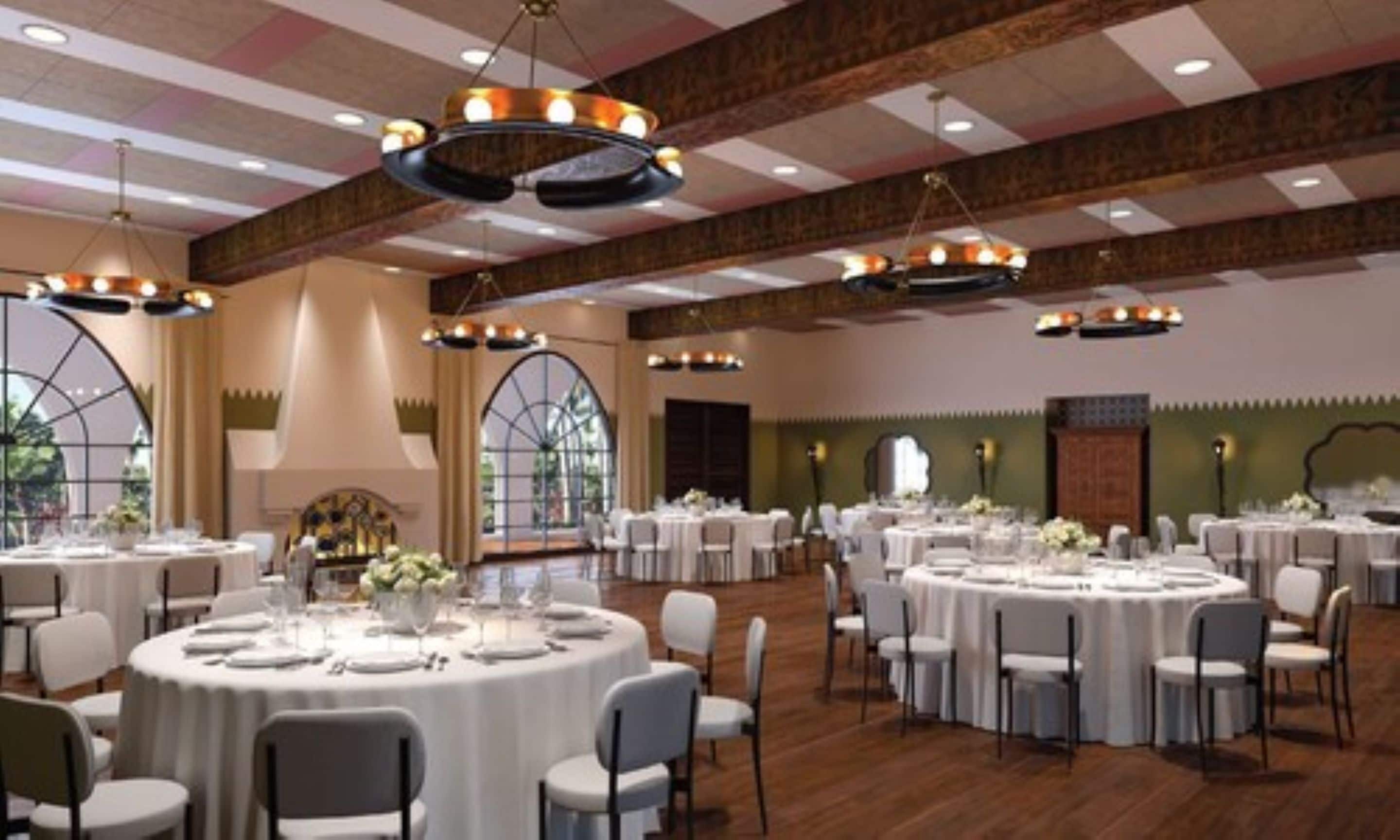 Une salle de banquet avec des tables rondes dressées pour le repas, de grandes fenêtres et un plafond haut avec des poutres apparentes.