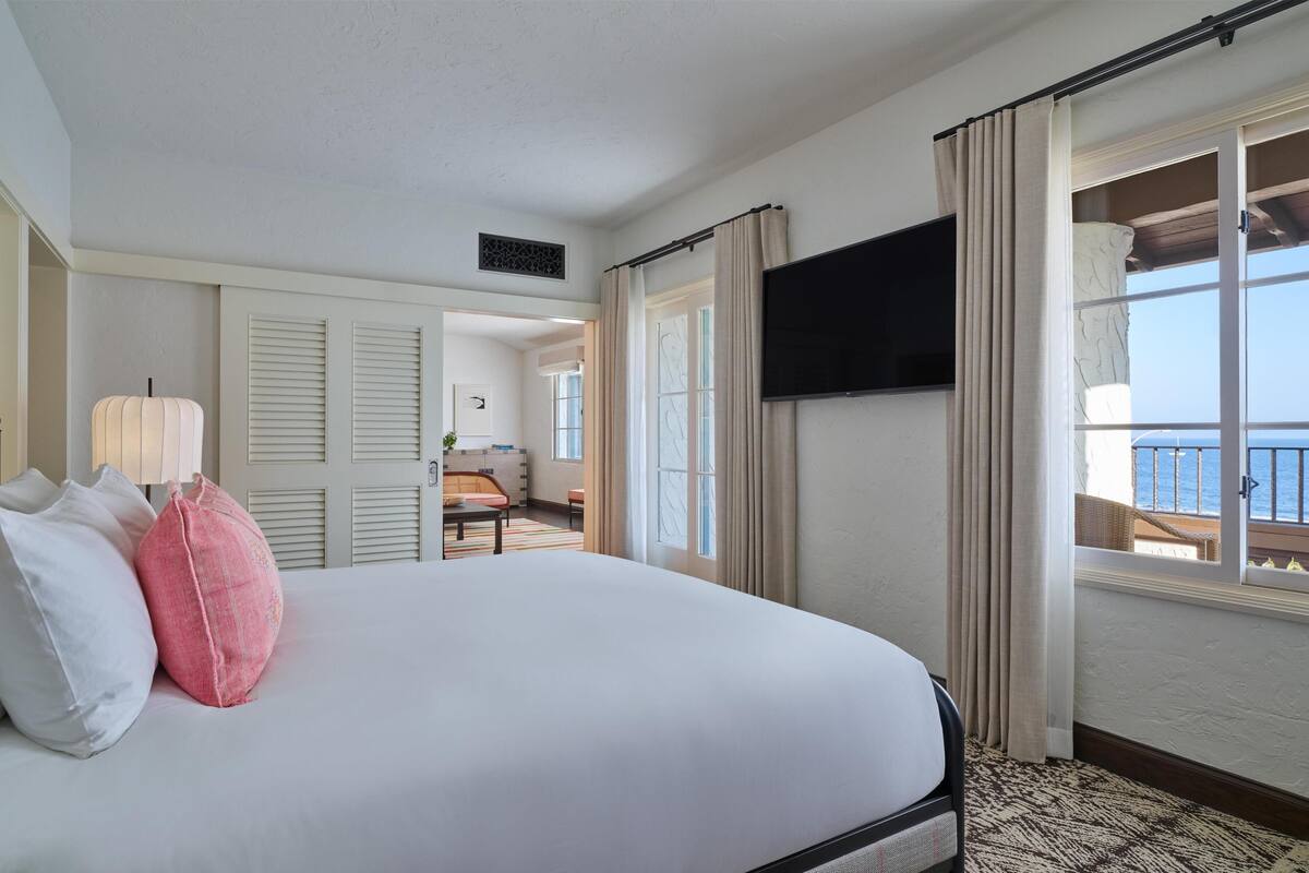 A hotel room with a bed, a television, a balcony with curtains, and a view of the sea.