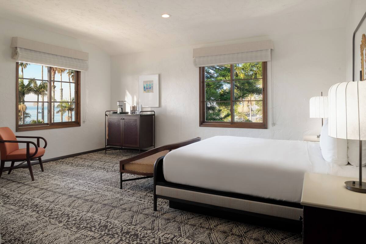 A hotel room with a bed, a desk, a chair, and a window with a view of palm trees and the sea.