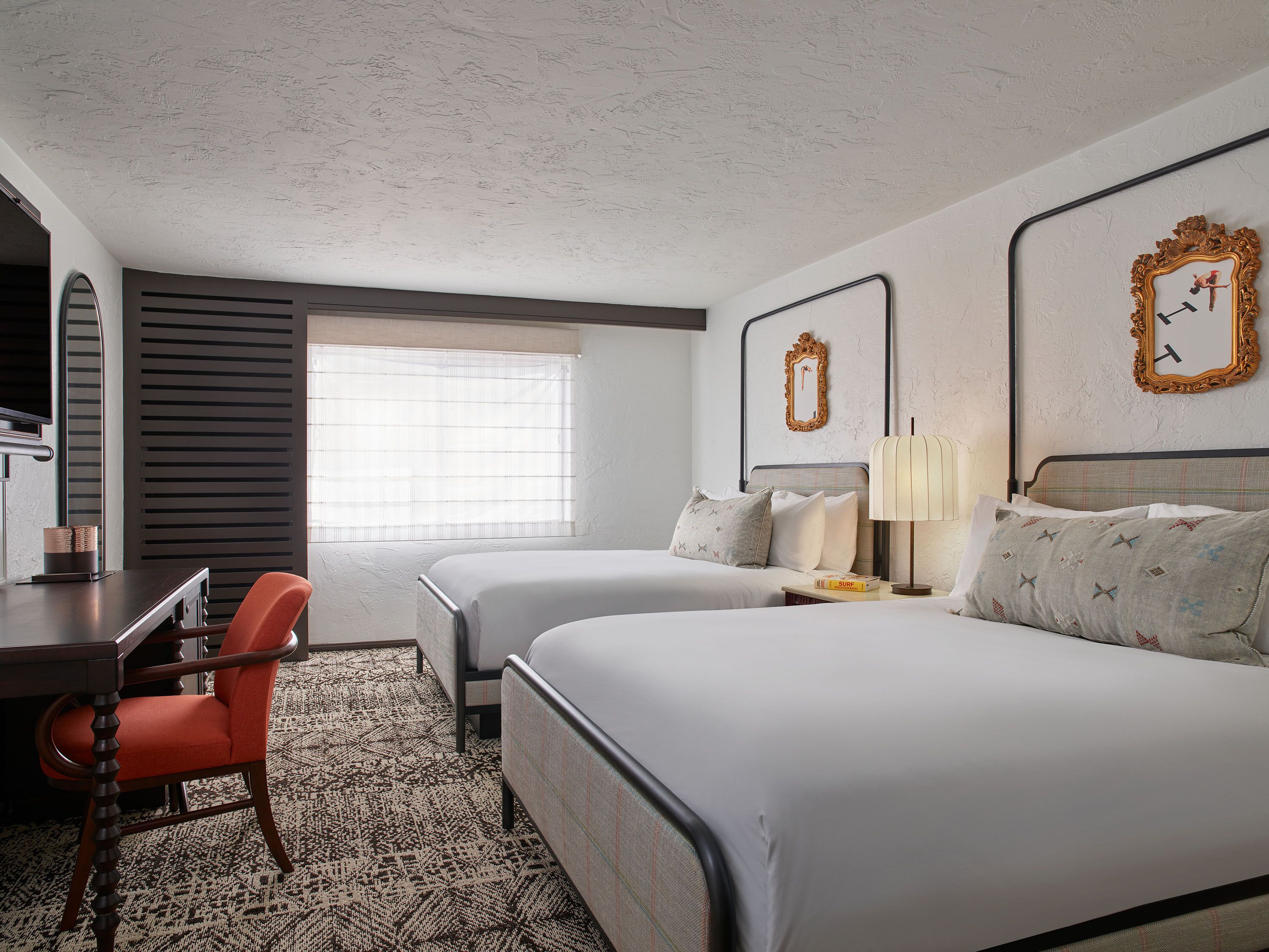 Habitación de hotel con dos camas, un escritorio y una silla. Cuenta con una ventana con persianas, un espejo en la pared y un reloj.